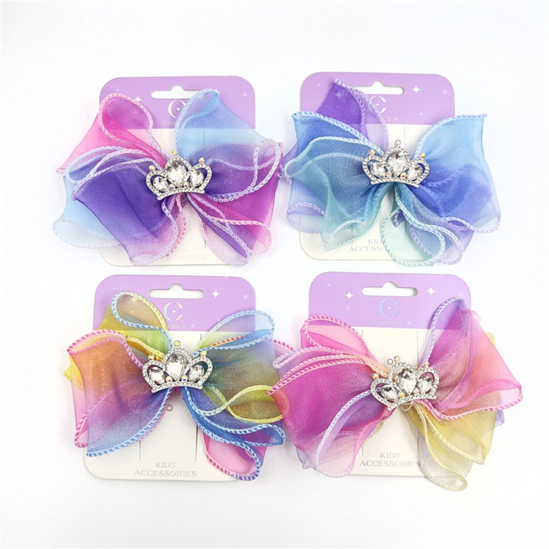 Large layered organza-style bow; Rainbow ombre colours with pink, blue, purple and yellow tones; Silver-tone crystal-look crown embellishment in the centre; Contrasting stitched edging on bow loops; Mounted on branded backing card labelled “Kids Accessories”;