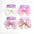 Large sheer organza bows in soft pink and white tones; Silver crown charm with small rhinestone-style details at the centre; Mounted on purple and cream backing cards labelled kids accessories; Wide, layered bow shape with gently curled edges;