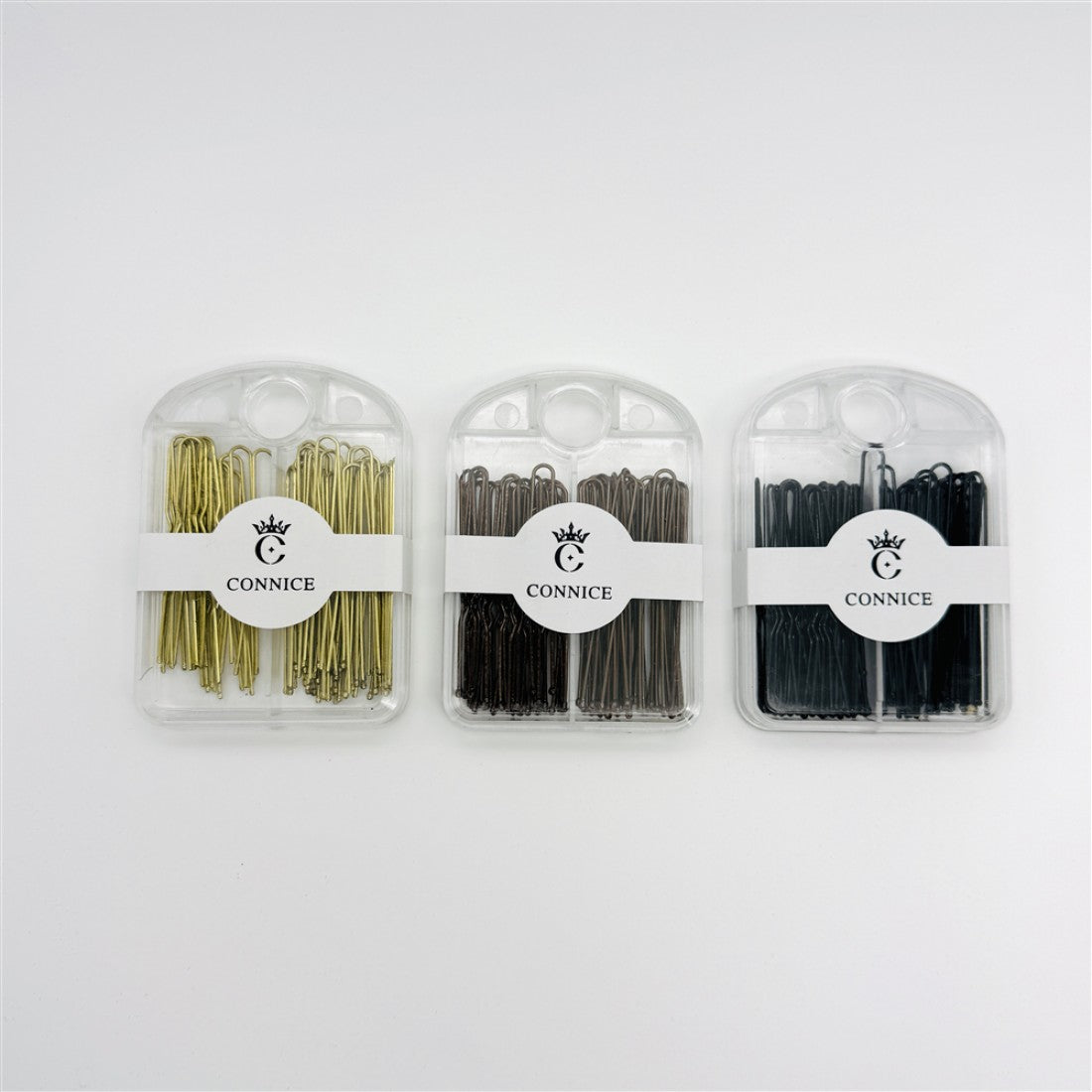 Three clear plastic cases shown side by side; Hair pins in gold, brown and black tones; Compact rectangular cases with rounded tops and hanging hole; White CONNICE branded bands wrapped around each case;