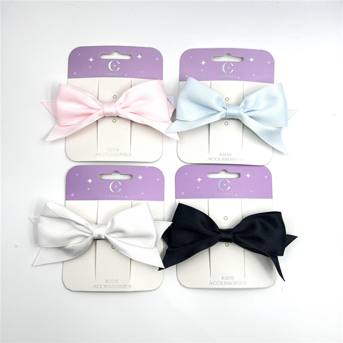 Large satin hair bows in pastel pink, light blue, white and black; Each bow has angled ribbon tails extending behind the main bow; Bows are presented on purple and white display cards; Packaging is branded as kids accessories;