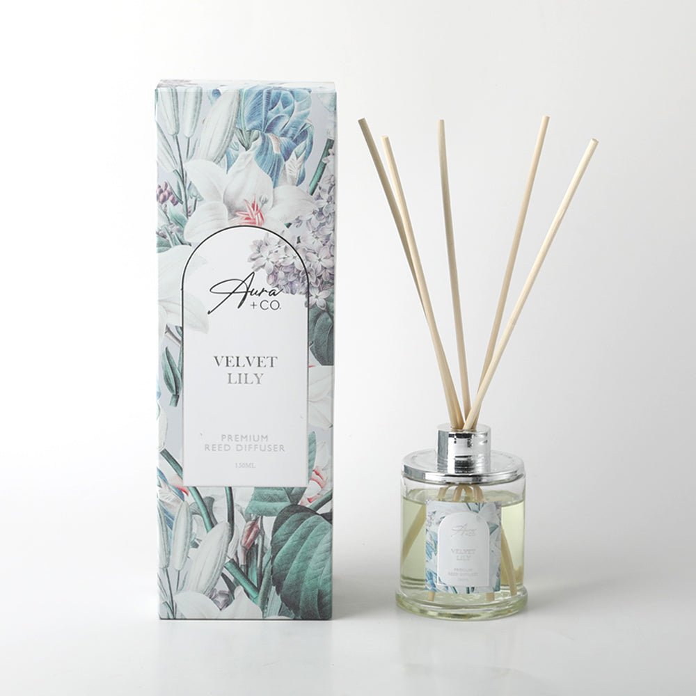 clear glass bottle with chrome top; natural wooden reeds; floral decorative box with white lilies and leaves; liquid diffuser visible inside bottle;