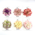 Large layered fabric flower on display card; Soft gradient colours in pastel tones; Full-bloom dahlia-style petal shape; Metal clip backing partly visible; Card backing printed with simple branding text;