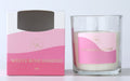 Candle in clear glass container; White wax candle; Pink label with gold text; Matching pink and white cardboard box packaging;